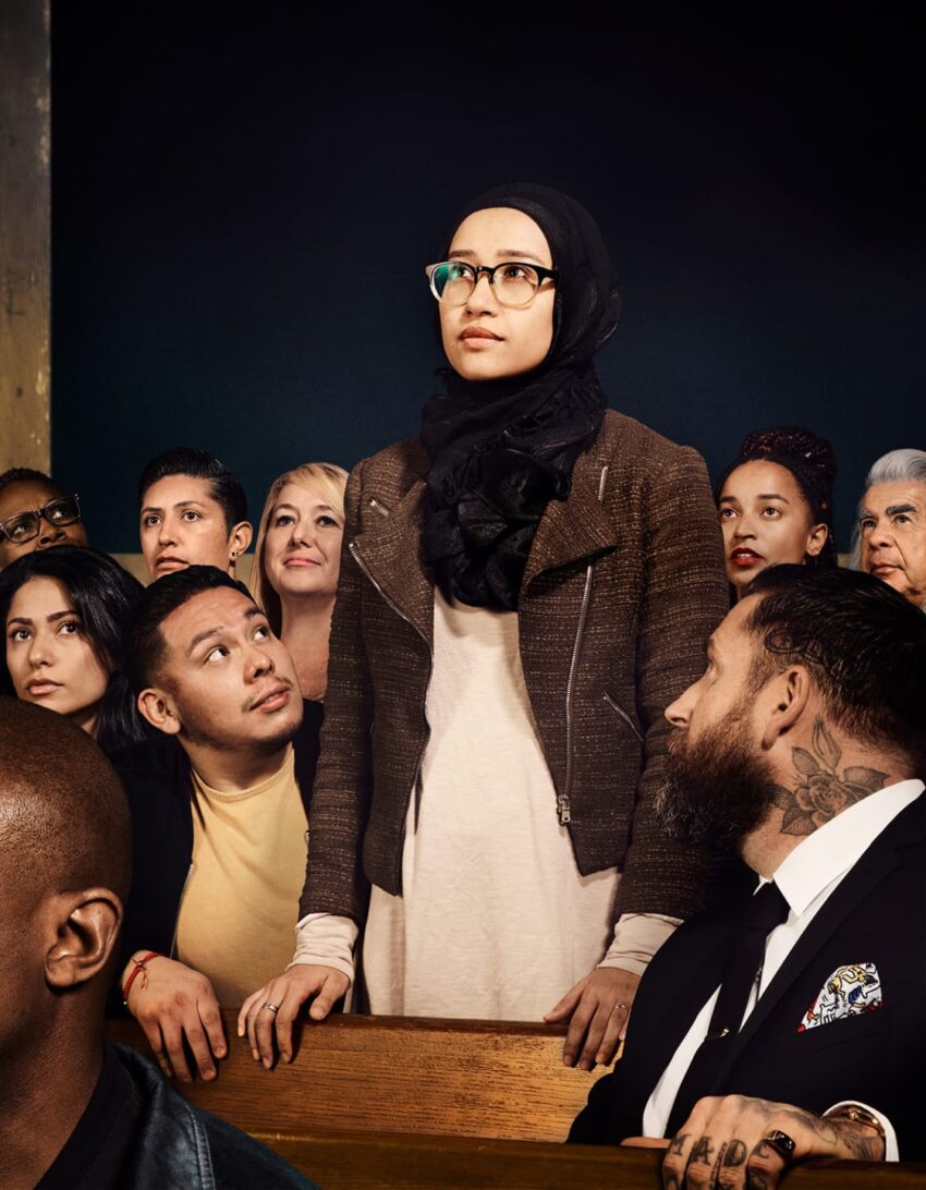 A diverse group of people in an auditorium, with a woman in a hijab standing, as others sit and listen attentively.