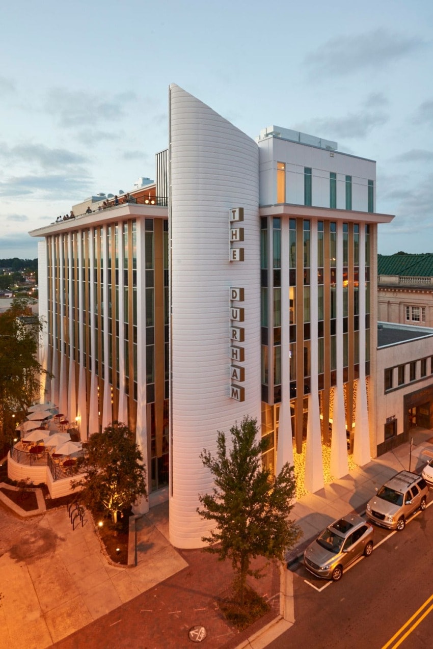 Modern hotel building with vertical windows and rooftop terrace at dusk, cars parked on street below.