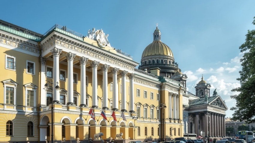 Historic building with columns in front, adorned with sculptures, alongside a domed structure under a clear blue sky.