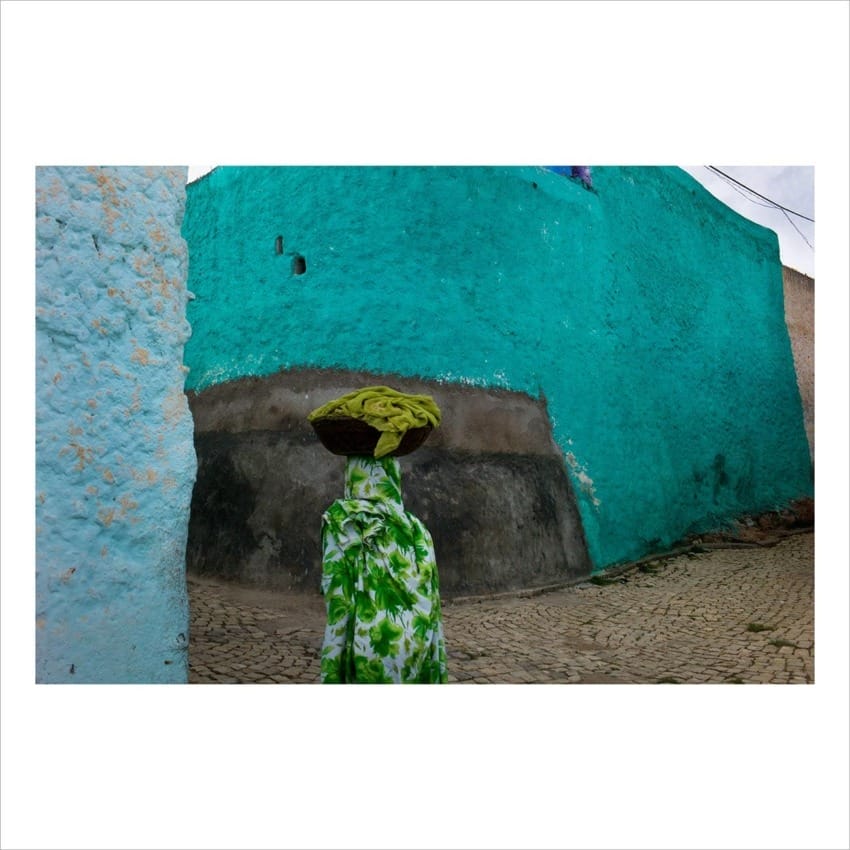 Person in patterned clothes carrying a bundle on head, walking on a cobblestone street with vibrant turquoise walls.