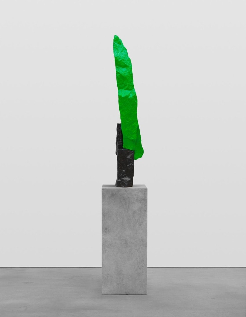 Modern abstract sculpture with a tall, slender green form atop a gray pedestal in a minimalistic gallery setting.