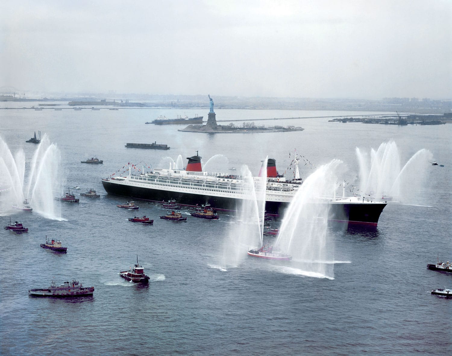 Ocean liner surrounded by water sprays from tugboats near the Statue of Liberty in New York Harbor.