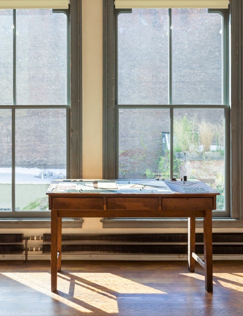 Sunlit wooden desk by large windows with papers, offering a view of greenery and a brick wall outside.
