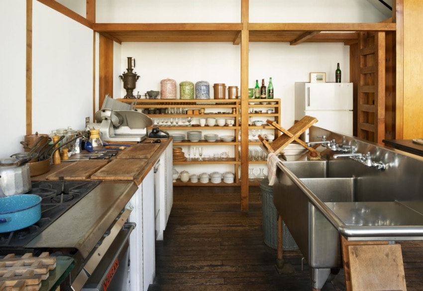 Rustic kitchen with wooden shelves, vintage cooking equipment, metal sink, and assorted dishes neatly arranged on shelves.