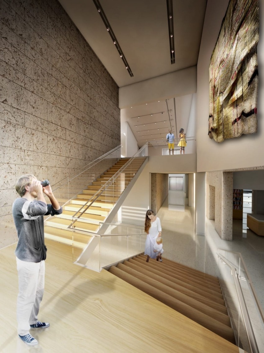 Visitor photographing an art installation in a modern museum with two people observing a wall textile from a balcony above.