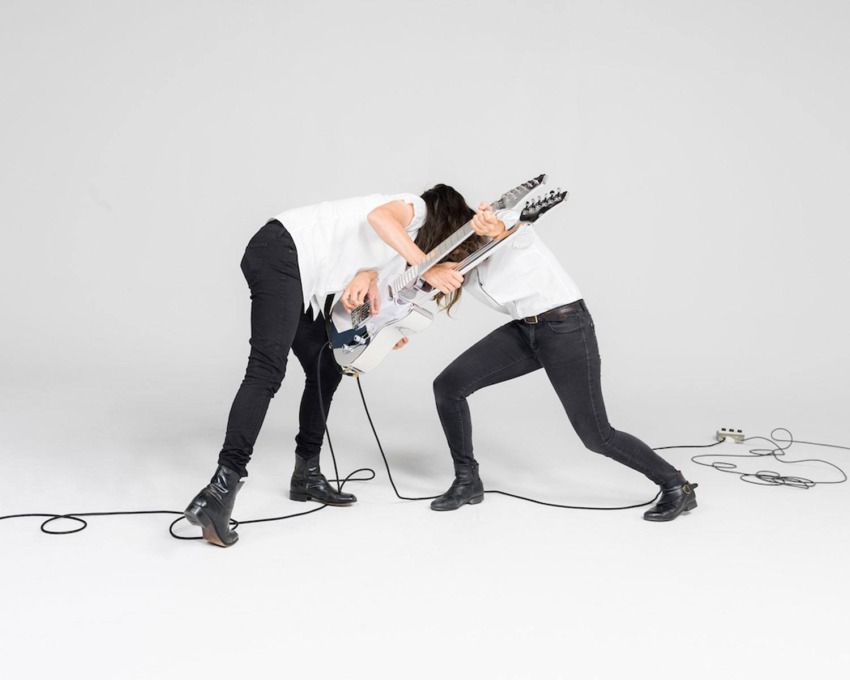 Two people in white shirts and black pants leaning against each other, each playing an electric guitar on a blank background.
