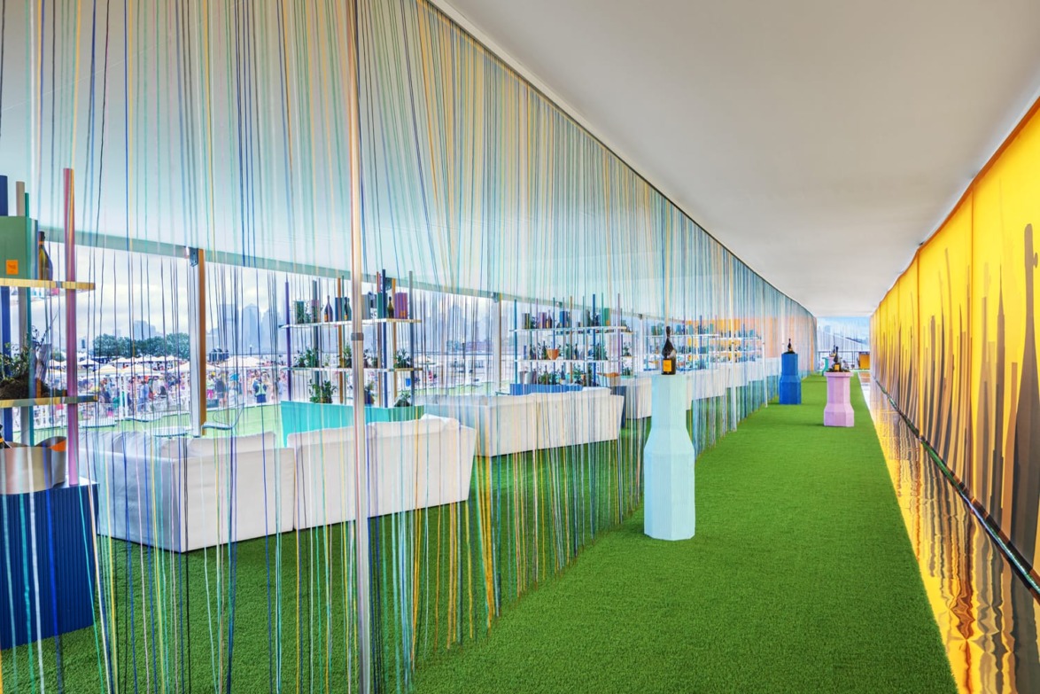 Colorful, modern lounge with green carpet, white furniture, and hanging strings, overlooking an outdoor event area.