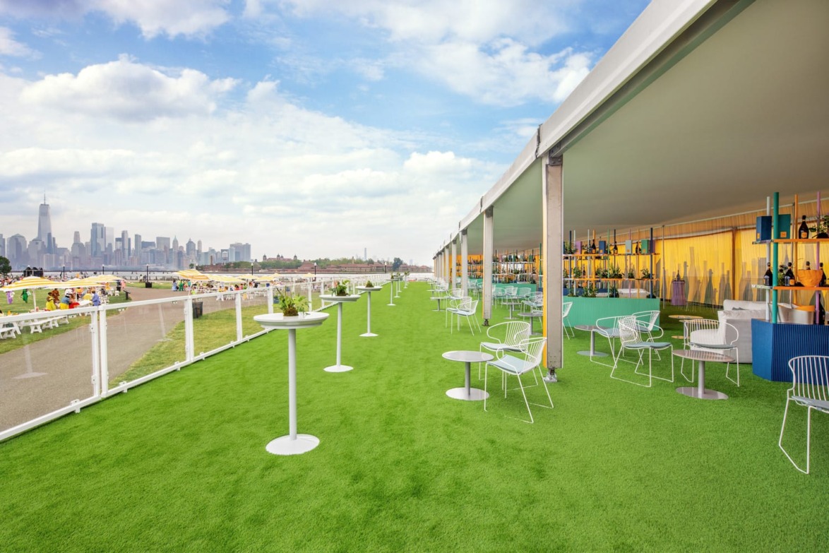 Outdoor event space with green artificial turf, tables, chairs, and a view of a city skyline under a partly cloudy sky.
