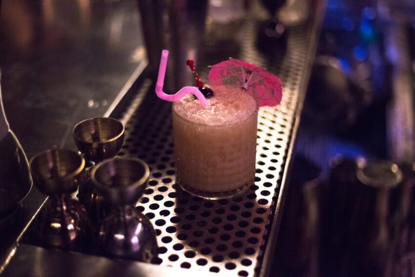 A pink cocktail with a curly straw and umbrella sitting on a perforated bar counter next to metal jiggers.