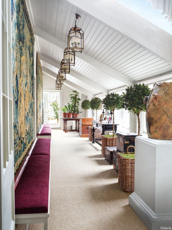 Sunlit hallway with white ceiling, tapestry wall, red bench, lantern lights, and decorative plants in a classic interior setting.