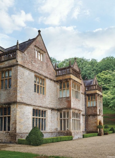 Historic stone mansion with large windows, ornate landscaping, and lush greenery under a blue sky with clouds.