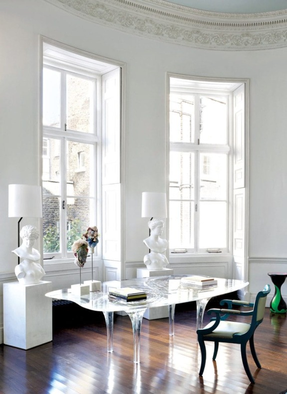 Elegant room with large windows, sculptural lamps, marble table, and teal chair on polished wooden floor.