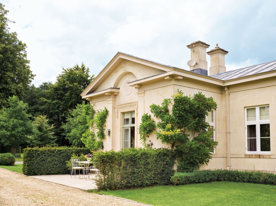 Elegant stone house with ivy, surrounded by lush greenery, small patio with chairs, and a gravel pathway in the foreground.