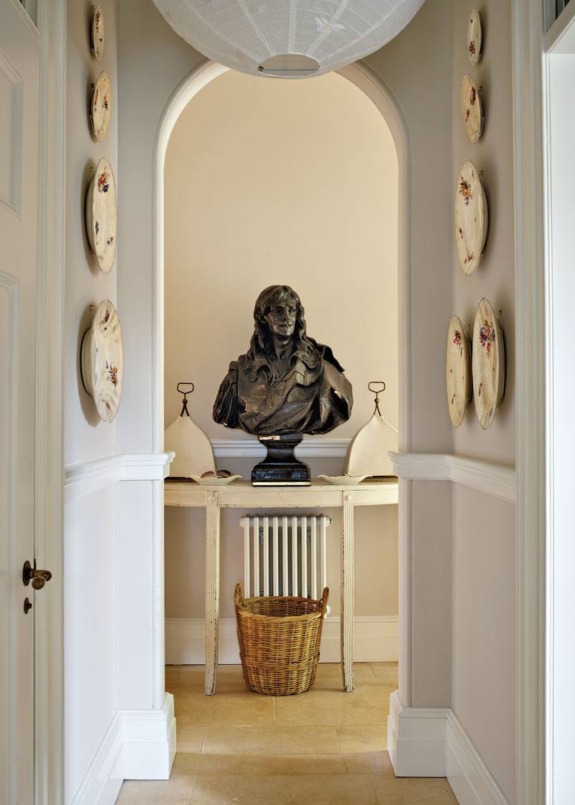 Elegant hallway with decorative plates on walls, classical bust on table, and wicker basket below in neutral-toned interior.
