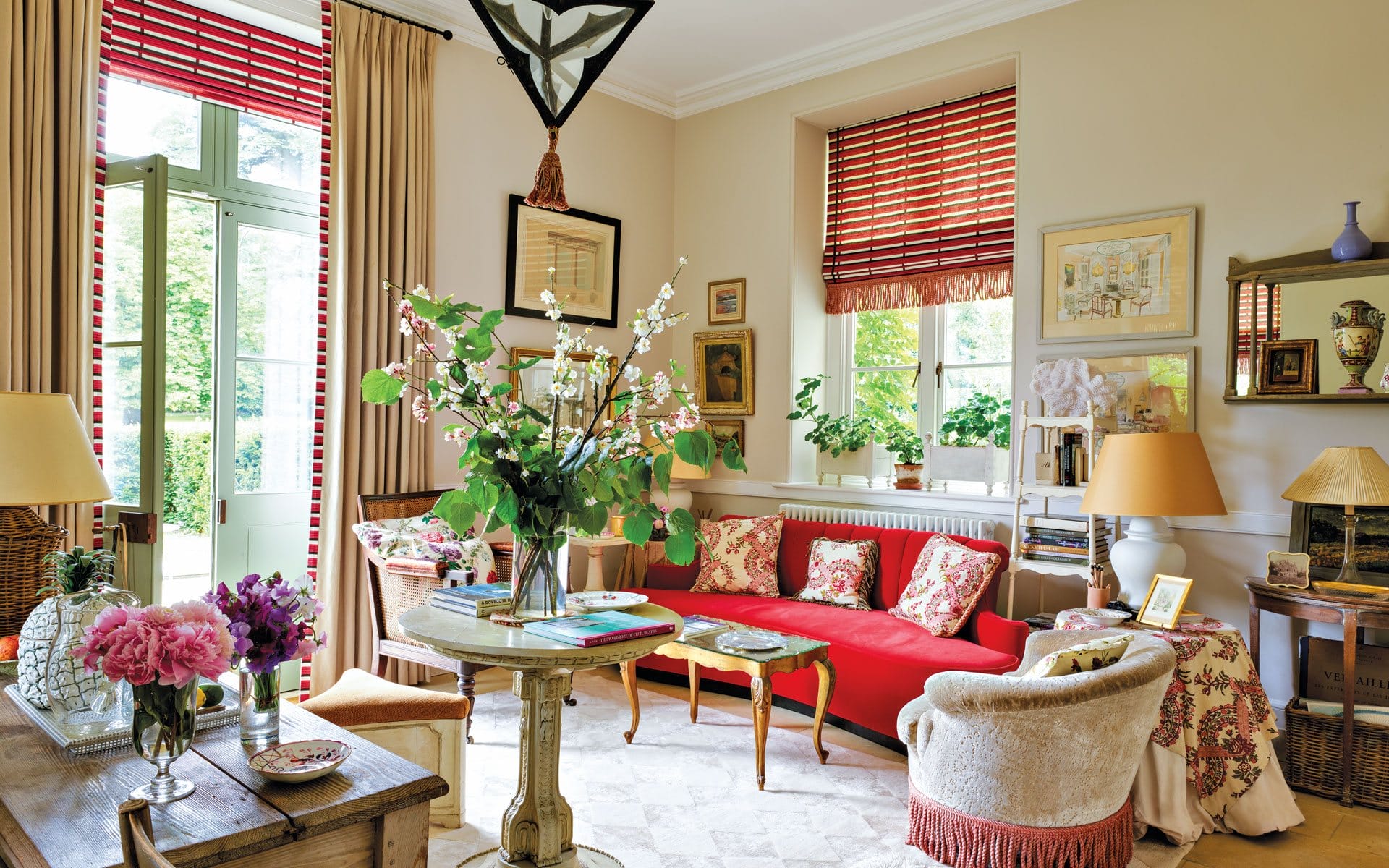 Colorful living room with red couch, plants, artwork, and large windows with red-striped blinds allowing natural light.