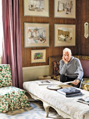 Elderly man sitting on a couch in a cozy living room with framed artwork and patterned furniture.