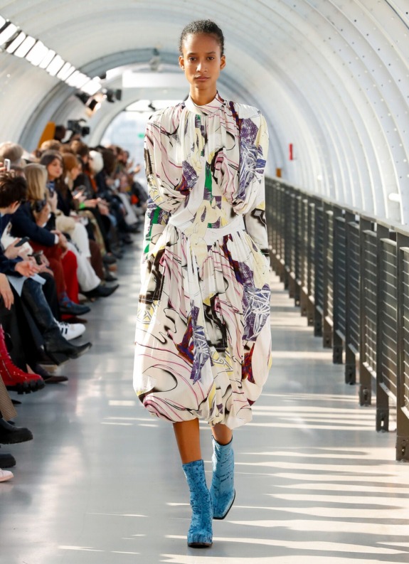 Model walking down a brightly lit fashion runway wearing a colorful abstract-patterned dress and blue textured boots.