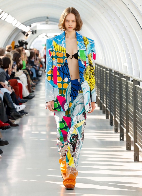 Model wearing colorful geometric suit walks runway in a modern, well-lit venue with seated audience.