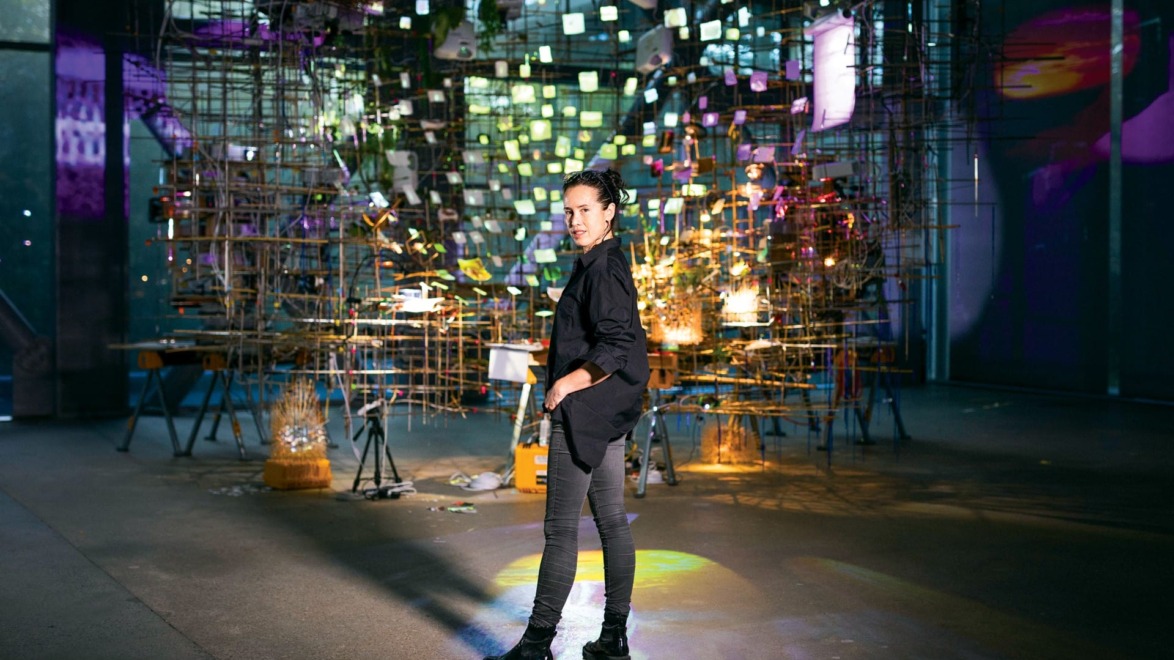 Artist stands in a dimly lit, vibrant art installation with colorful lights and intricate structures in the background.