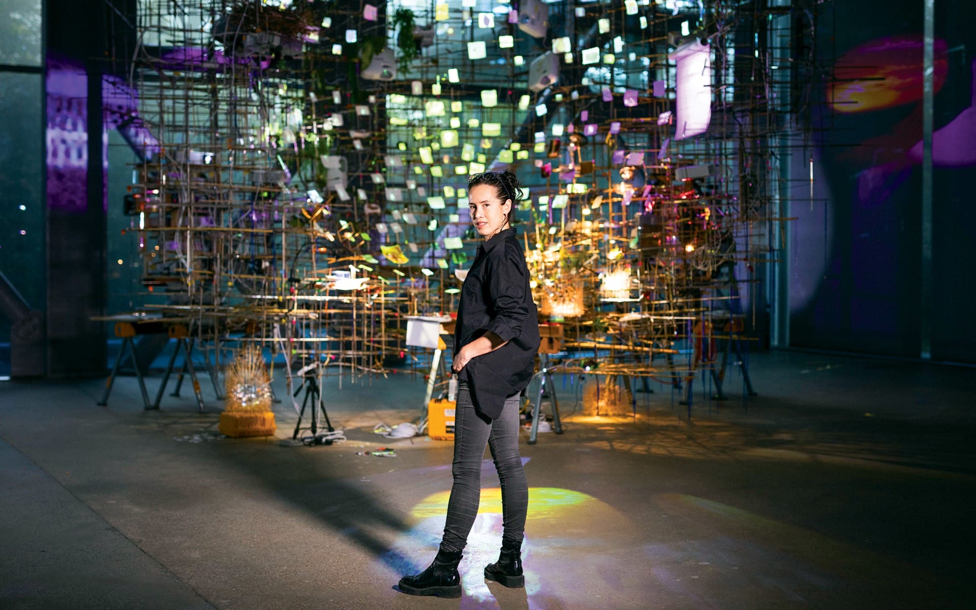 Artist stands in a dimly lit, vibrant art installation with colorful lights and intricate structures in the background.