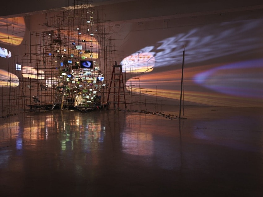 Art installation with screens on scaffolding, creating light reflections on a polished floor in a dimly lit gallery space.