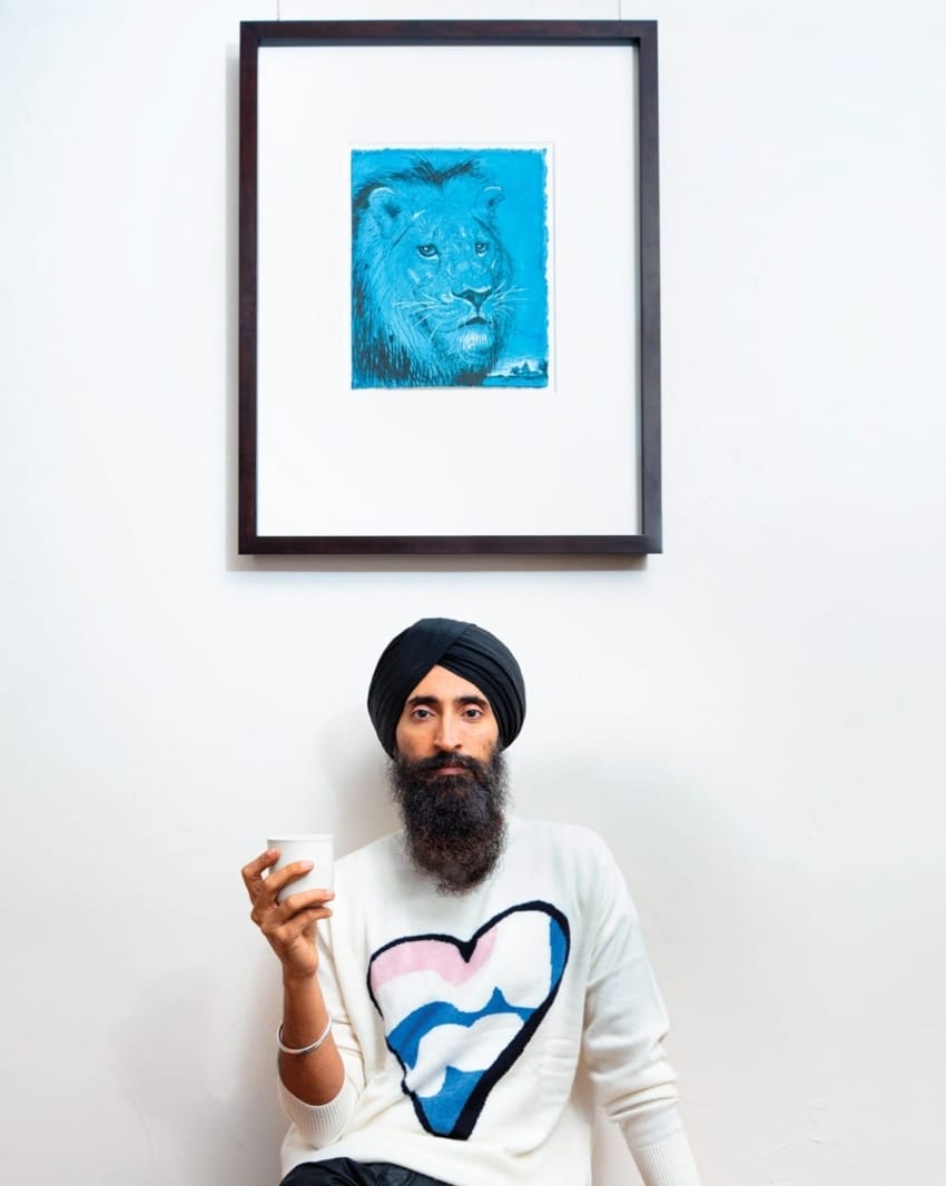 Man in a turban holding a cup, sitting under a framed blue lion drawing on a wall.