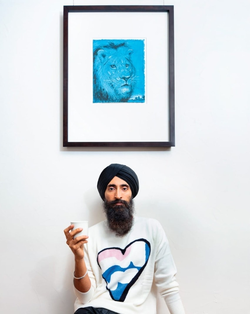 Man in a turban holding a cup, sitting under a framed blue lion drawing on a wall.