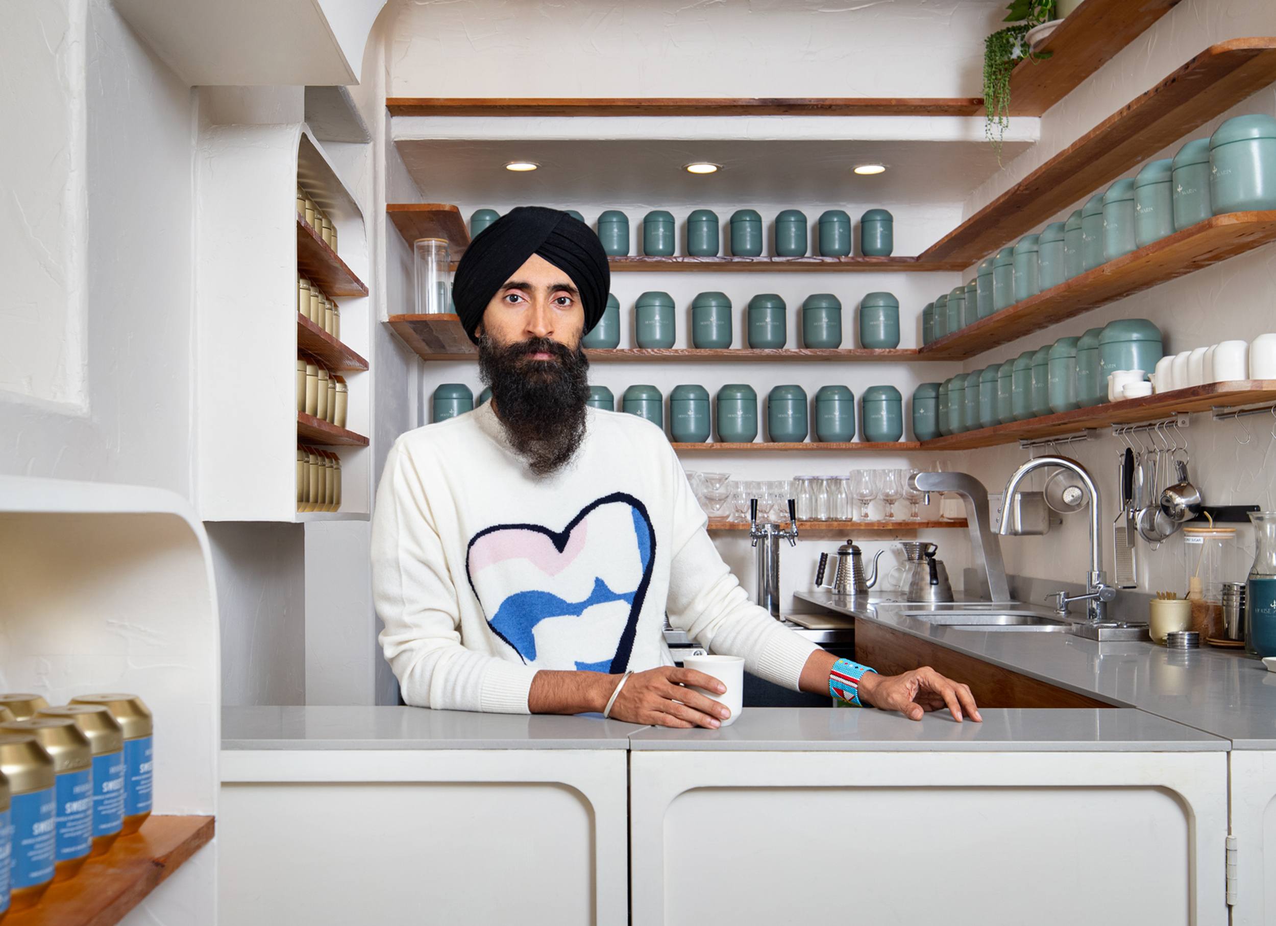Person with a beard and turban standing behind a counter in a modern kitchen or shop, surrounded by jars and shelves.