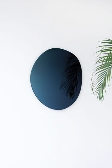 Round black wall mirror with a reflection of palm leaves, mounted on a white wall.