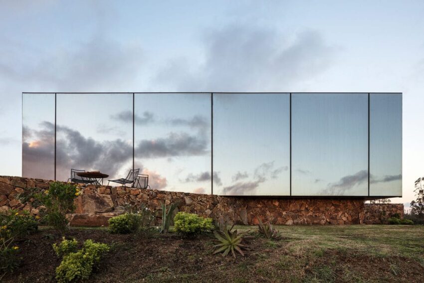 Modern building with mirrored walls reflecting cloudy sky, set on a stone base with surrounding greenery at dusk.