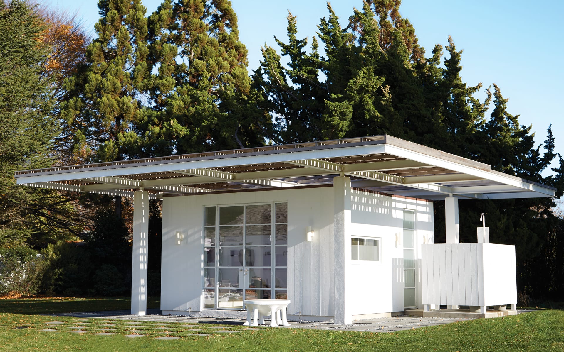 Modern white tiny house with large windows, flat roof, and surrounded by lush green trees on a sunny day.
