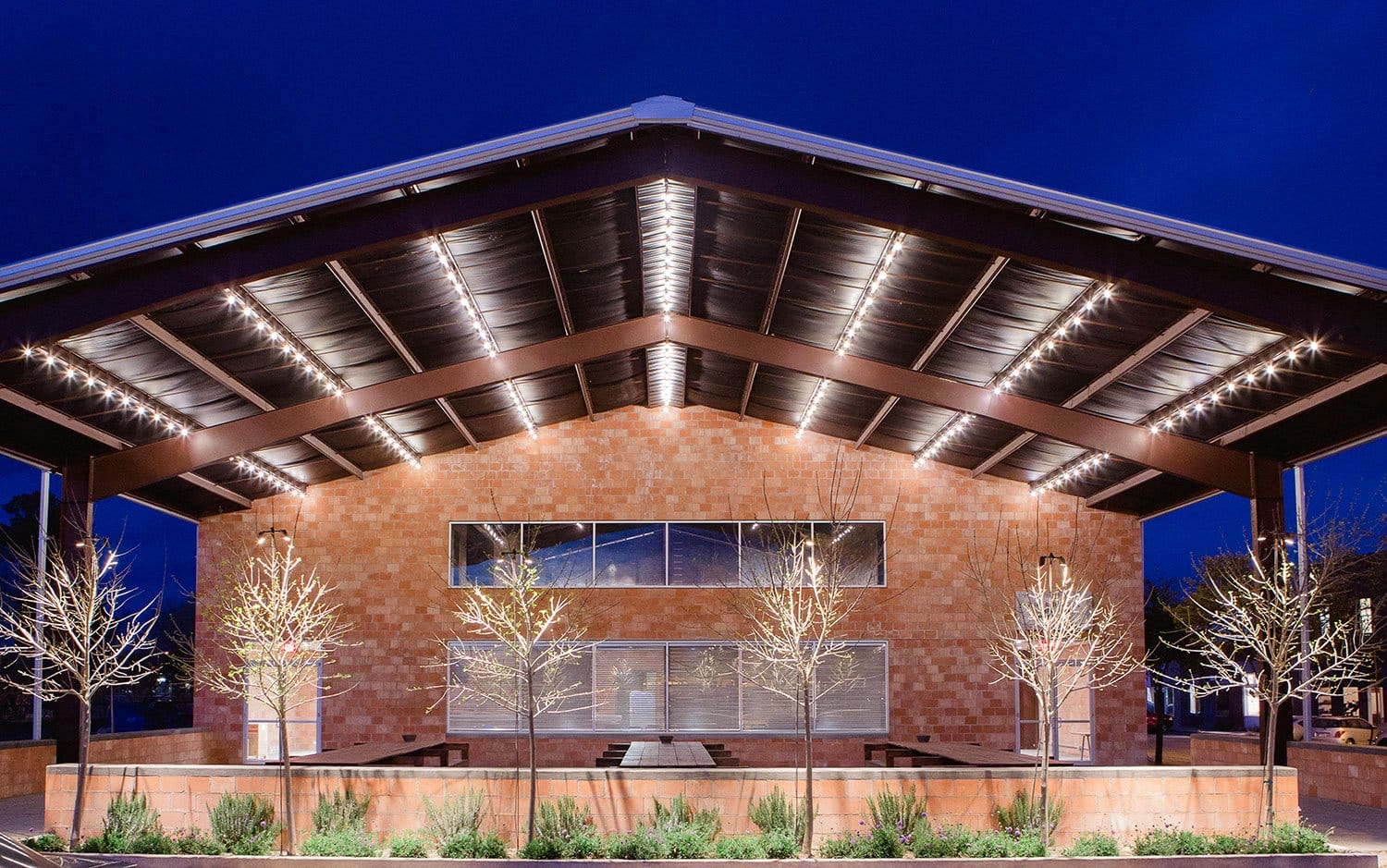 Modern building exterior with illuminated roof at night, featuring trees and landscaped greenery in the foreground.