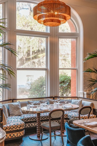 Cozy restaurant corner with a set table, surrounded by tall windows and plants, under a wicker light fixture.