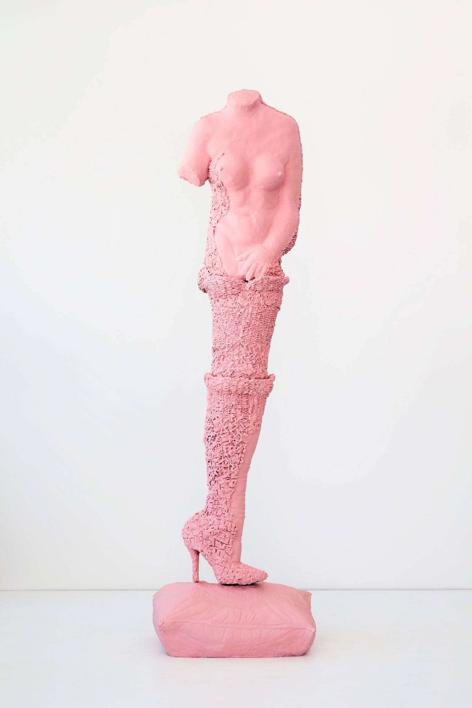 Contemporary sculpture of a pink female torso with one leg, wearing high-heeled boot, on a pedestal against white background.