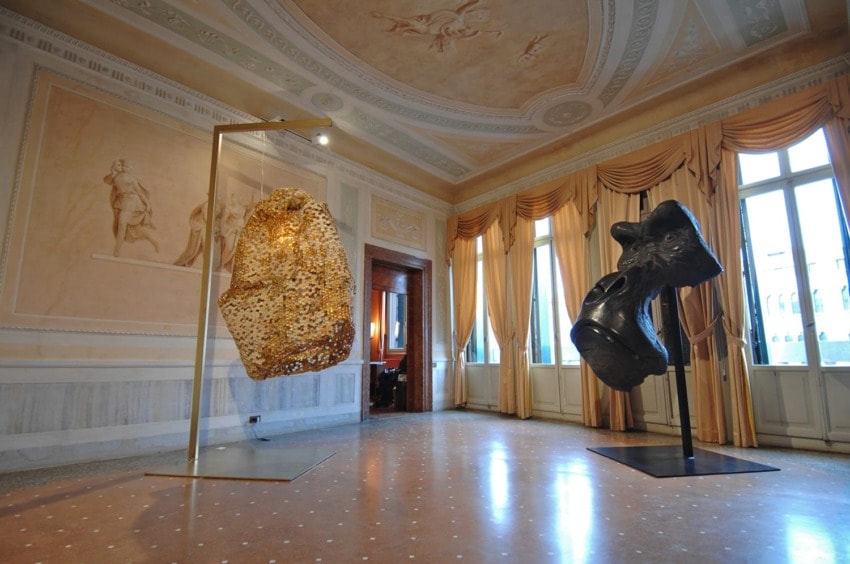 Two abstract sculptures displayed in an ornate room with tall windows and decorative ceiling, casting shadows on the floor.