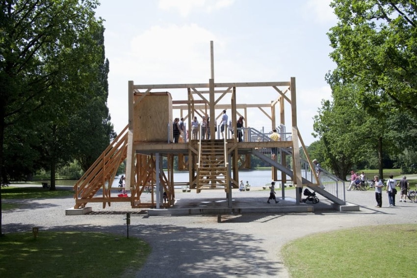 Wooden playground structure with children playing on it in a park surrounded by green trees and a clear blue sky.