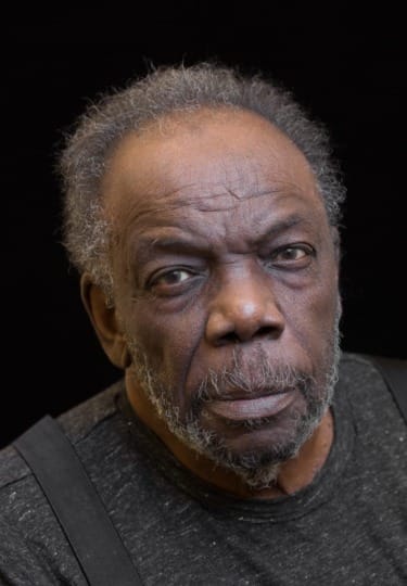 Elderly man with a contemplative expression, wearing a grey shirt and suspenders against a black background.
