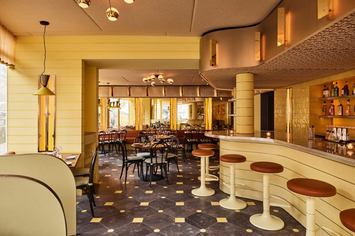 Art Deco-inspired cafe interior with yellow walls, round bar, bar stools, and dark checkered floor.