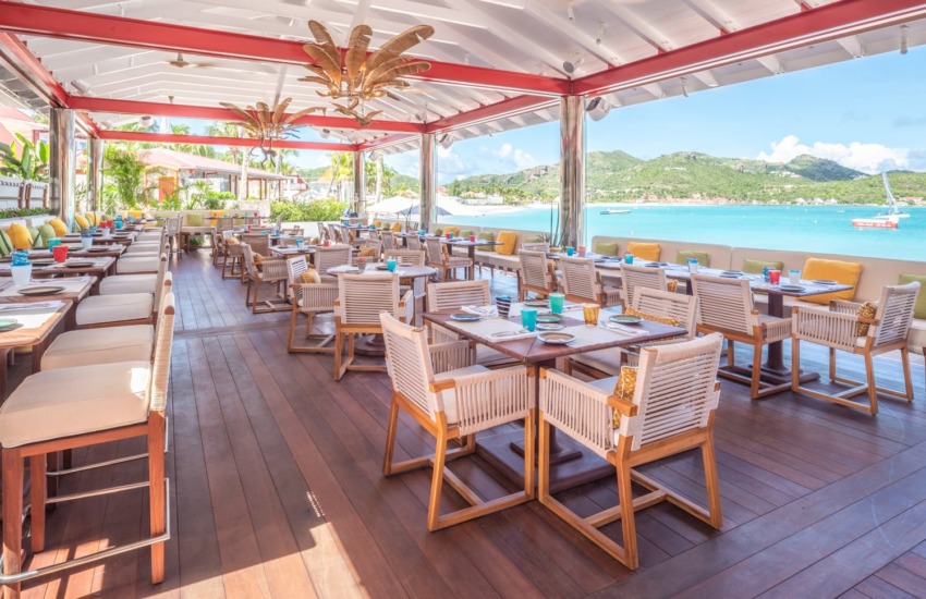 Spacious outdoor restaurant with wooden tables, chairs, and ocean view in a tropical setting under a white and red roof.