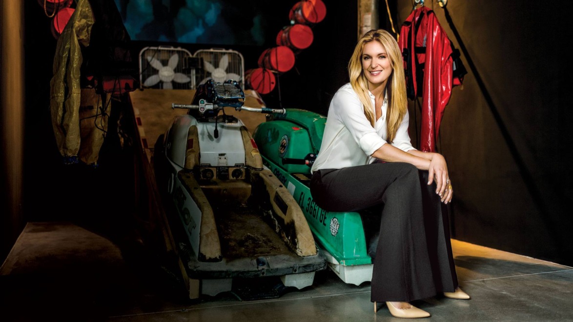 Woman sitting on vintage snowmobile in a stylish room filled with colorful jackets and unique industrial decor.