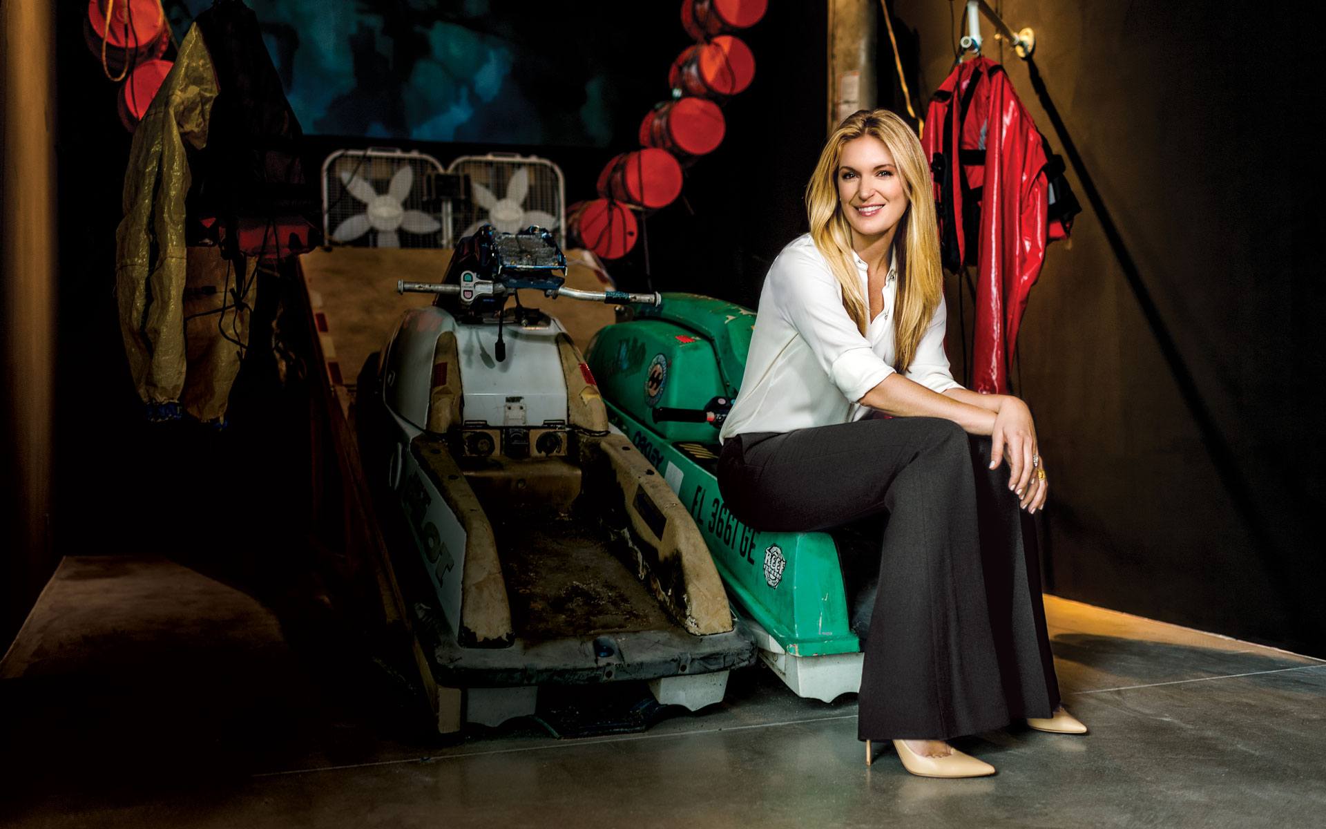 Woman sitting on vintage snowmobile in a stylish room filled with colorful jackets and unique industrial decor.