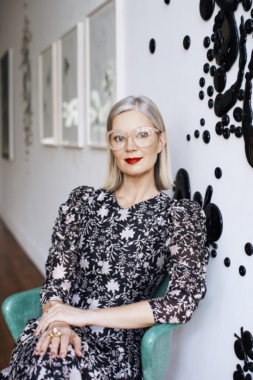 Person with glasses in patterned dress sitting on green chair, in art gallery with abstract wall decorations.
