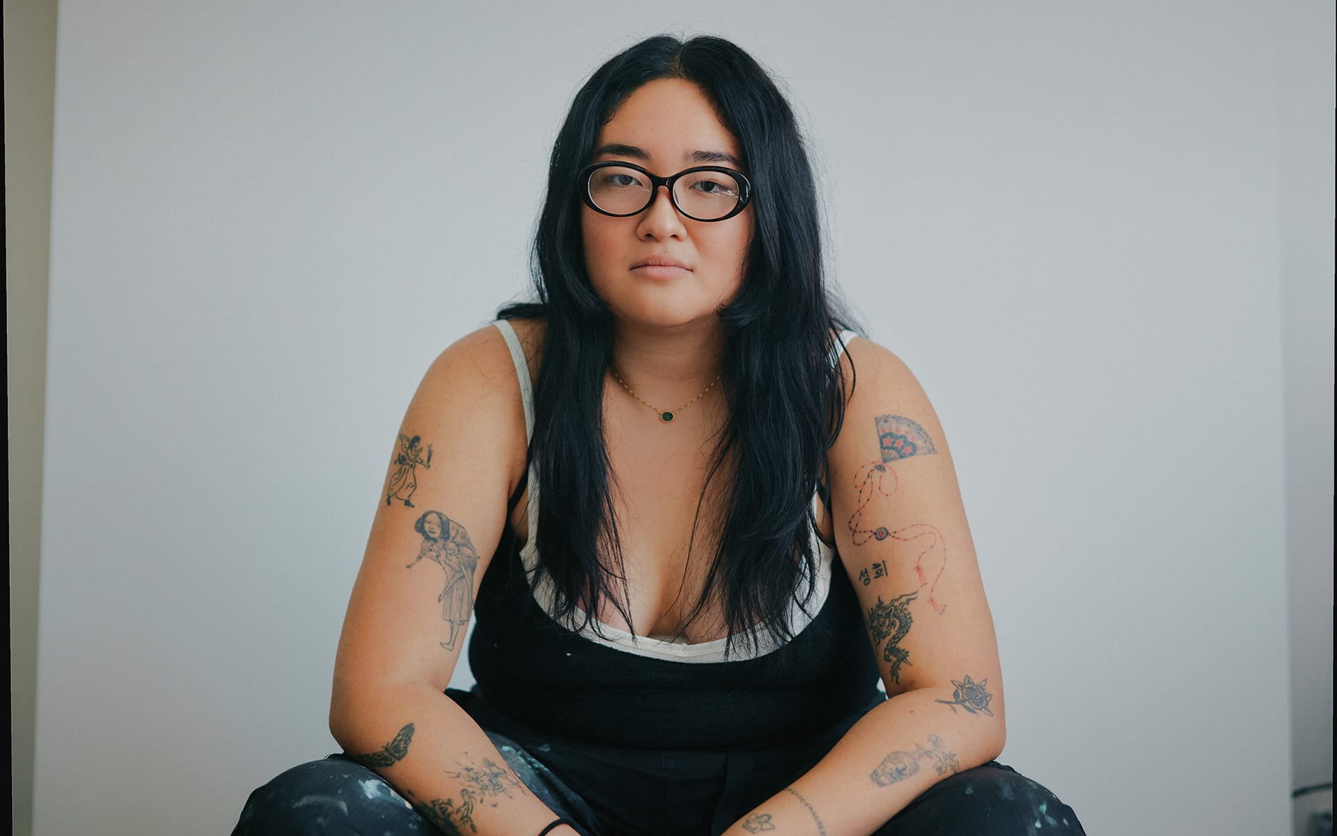 Person with long hair and glasses, seated, wearing a black and white top and displaying various tattoos on arms.