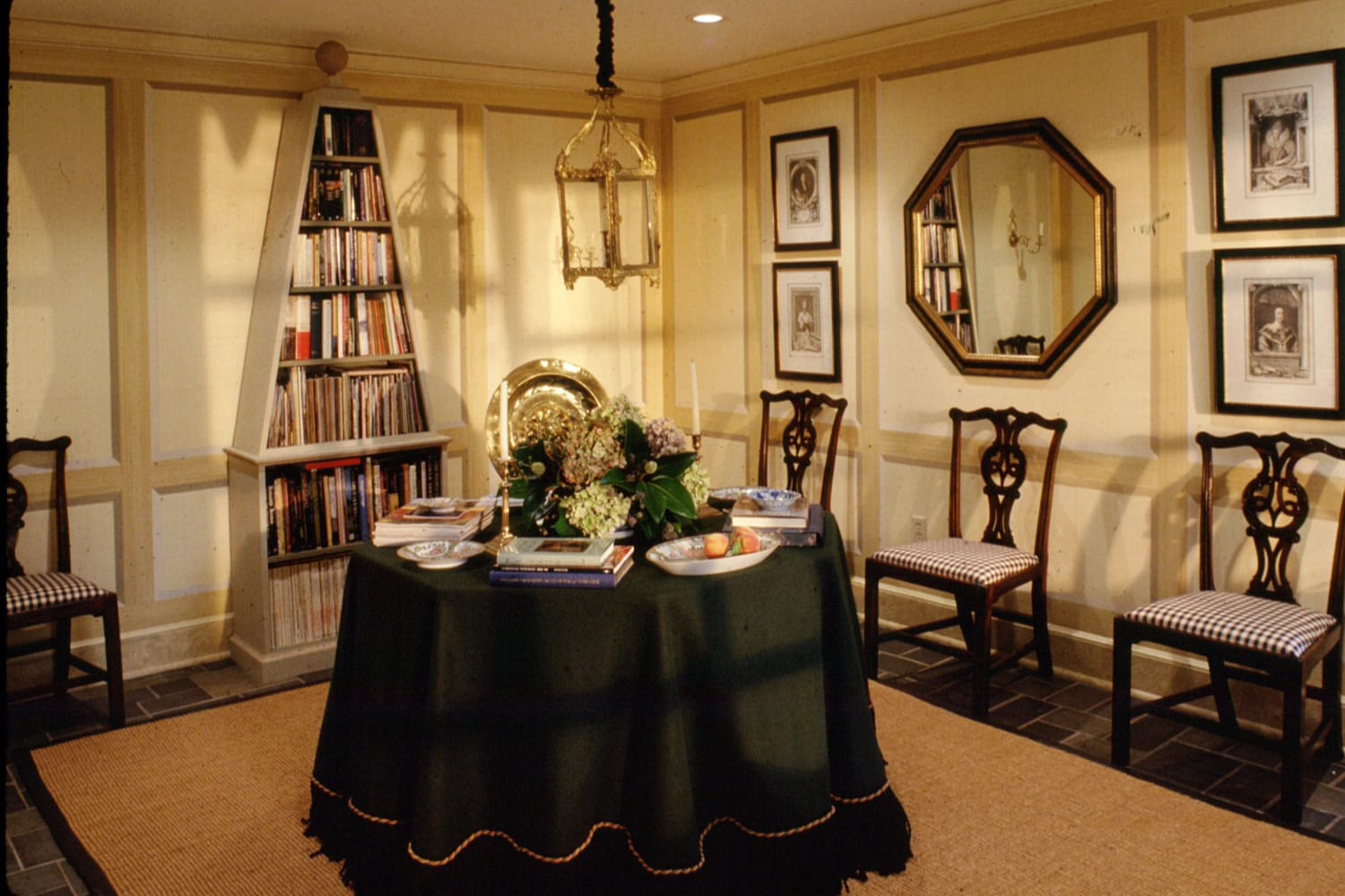 Dining room with round table, bookshelves, and art on walls, featuring a green tablecloth and chandelier lighting.