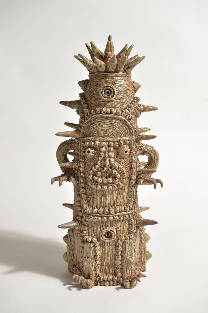 Ceramic sculpture with intricate carved details, featuring a face and multiple protruding spikes, on a plain background.