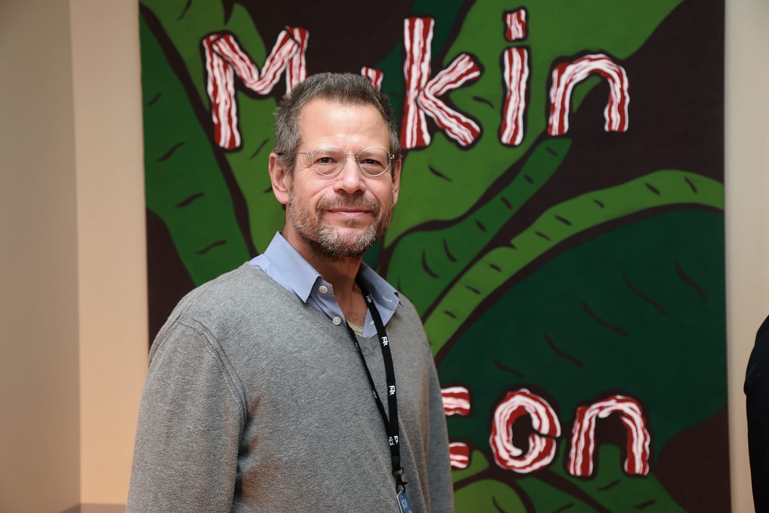 Person standing in front of a colorful mural with abstract shapes and letters, wearing glasses and a gray sweater.