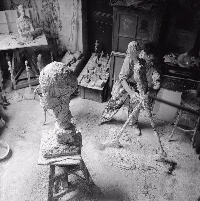Artist working on sculptures in a cluttered studio with clay figures on pedestals and tools scattered around.