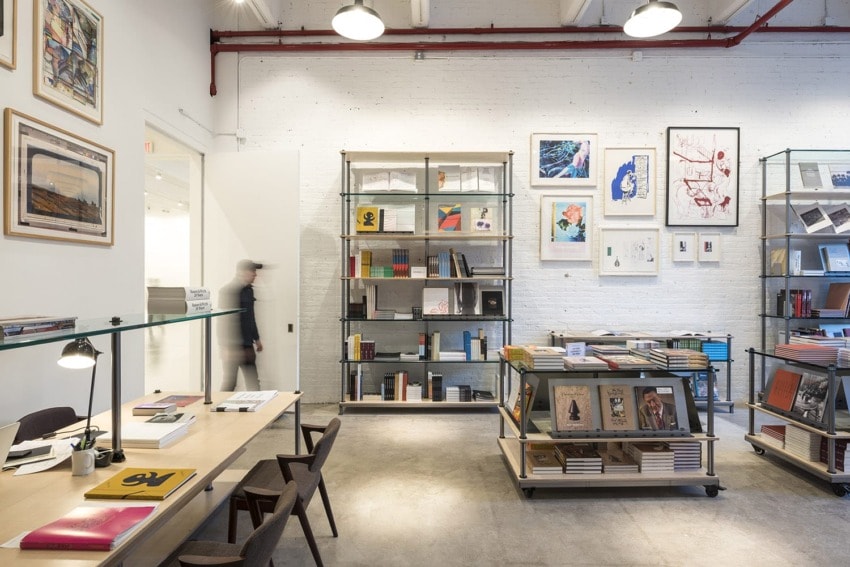 Modern bookstore interior with wooden tables, chairs, shelves filled with books and artwork on white brick walls.