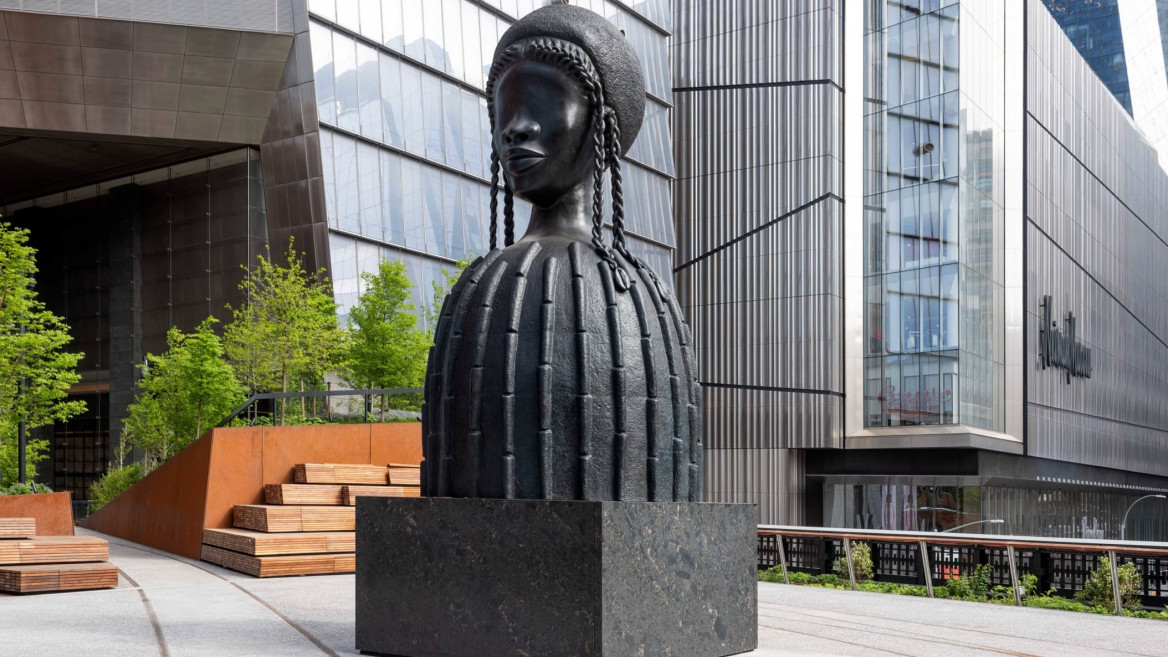 Sculpture of a large bust of a woman wearing a helmet with braids in an urban setting with modern buildings in the background.
