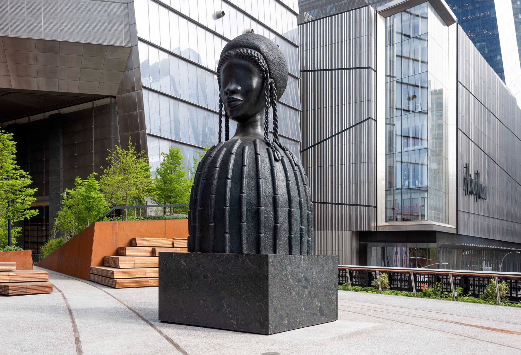 Sculpture of a large bust of a woman wearing a helmet with braids in an urban setting with modern buildings in the background.